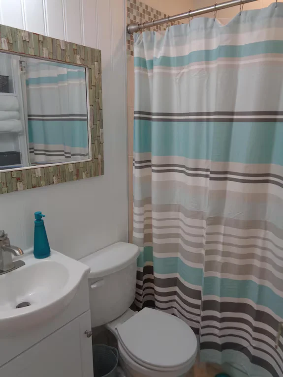 Small bathroom before remodel with white vanity, toilet, striped blue shower curtain, and wall mirror in an Outer Banks home.